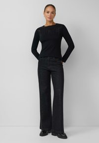 Schwarzes Langarmoberteil mit Strassverzierungen auf der Schulterpartie, kombiniert mit dunklen Schlagjeans und schwarzen Plateauschuhen. Einfarbiger Hintergrund.