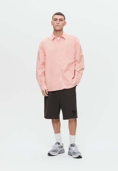 Young man standing wearing a pink and white striped button-up shirt, black knee-length shorts, white socks, and gray sneakers.