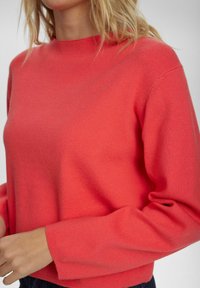 Koralo spalvos megztinis su ilgomis rankovėmis, apvaliu kaklu ir tekstūruotu paviršiumi. Apatinė dalis šiek tiek trumpesnė, prigludusi, su minimaliais siuvinėjimo elementais.