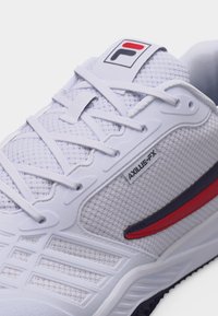 Λευκό αθλητικό παπούτσι με επάνω μέρος από δίχτυ, λευκά κορδόνια, ετικέτα "AXILUS-FX", κόκκινη και ναυτική καμπυλωτή ρίγα, και λογότυπο FILA στη γλώσσα.