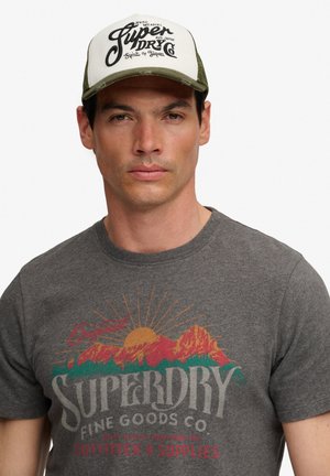 Man draagt een grijs Superdry T-shirt met bergafbeelding en een witte en groene Superdry pet die naar voren kijkt.