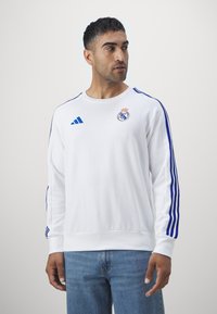 Fehér pulóver kék csíkokkal, kerek nyakkal és bordázott mandzsettákkal. A mellkason látható a Real Madrid logója és az Adidas márkajelzés.