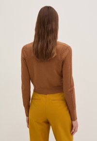 Suéter marrón de punto con un diseño recortado y mangas largas, que presenta detalles de cortes verticales a lo largo de los hombros, combinado con pantalones amarillos.