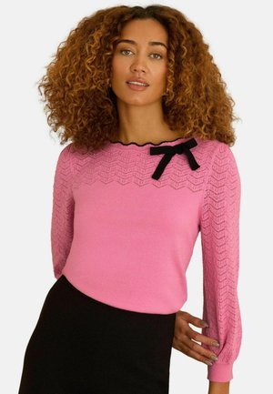 Moterys su garbanotais plaukais, dėvinčios rožinį megztinį su nėrinių detalėmis ir juodu kaspinu ant krūtinės, derintą su juoda sijonu.