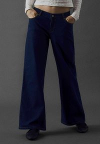 Breda mörkblå jeans med en slät textur, som har framfickor och knappstängning, i kombination med svarta platta skor.