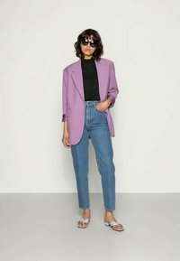 Blazer oversized lavanda sobre uma blusa preta, combinado com jeans azul claro e sandálias brancas. Os acessórios incluem óculos de sol.