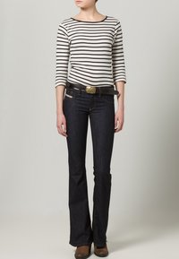 W paski bluzka z długim rękawem w kolorze kremowym i czarnym, zestawiona z ciemnoniebieskimi dżinsami z rozszerzanymi nogawkami. Brązowy pasek z złotą klamrą i brązowe buty.