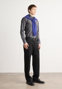 Fekete nadrág, szürke ing és kék pulóver a vállakon. Simaságú anyag, egyenes szabás, öv akcentussal, fekete cipő. Minimalista design.