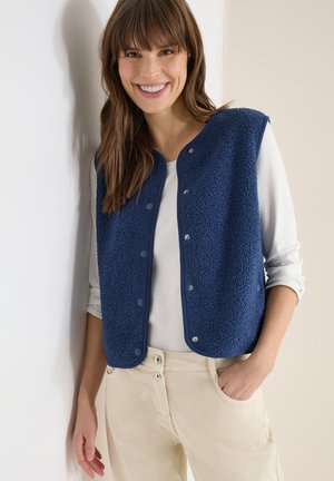 Femme souriante portant une veste polaire bleu marine à boutons par-dessus une chemise blanche à manches longues, debout avec une main dans la poche de son pantalon beige.