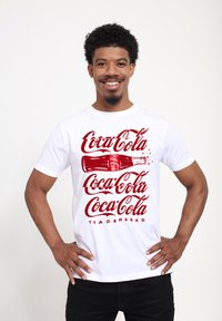 Bílý bavlněný tričko s červeným logem Coca-Cola a ilustrací lahve, doplněné opakovaným názvem značky v zakřiveném písmu.