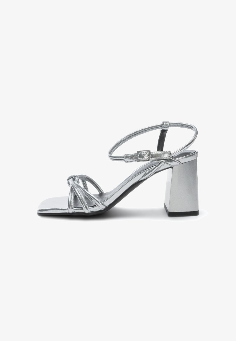 Hopeaiset korkokengät suoralla varpaalla, metalliset hihnat, tasainen nilkkahihna ja paksu korko. Sujuva pinta ja heijastava pinta.