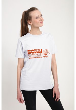 Ung kvinde med brunt hestehale ser op, iført en hvid T-shirt med orange "Soul 1 Skateboards Los Angeles County" grafik og sorte bukser.