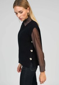 Pull en maille noir avec un col en V, agrémenté de boutons sur le côté. Porté par-dessus une blouse longue à manches transparentes marron. Un jean noir complète la tenue.