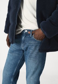 Chaqueta de forro polar azul marino sobre una camisa blanca, acompañada de unos jeans de mezclilla azules. Los jeans tienen un cierre de botón y un ligero desgaste.