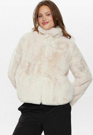 Veste en fausse fourrure de couleur beige clair avec un col haut, une forme carrée, une texture douce et une fermeture à boutons ; associée à un pantalon noir.