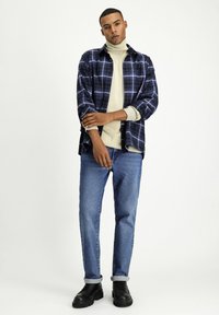 Camicia a scacchi blu e tartan, dolcevita crema, jeans azzurri chiari, risvoltini, scarpe di pelle nere. Il modello posa con le braccia incrociate.