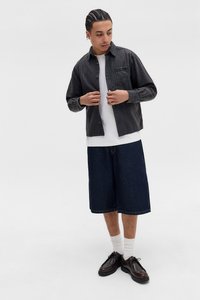 Svart og grå rutete skjorte med knapper over en hvit t-skjorte, kombinert med knelengde mørk denimshorts og svarte sko.