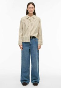 Chemise beige à rayures boutonnée avec une coupe décontractée, associée à un jean large bleu clair et des chaussures marron. Design simple avec des rayures contrastantes.