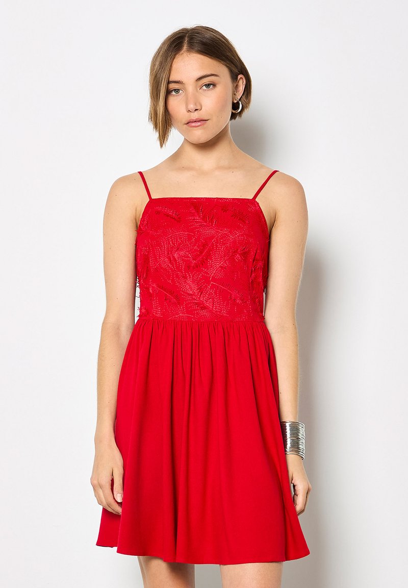 Jeune femme portant une robe rouge sans manches avec un corsage brodé et une jupe plissée, debout devant un fond blanc uni.