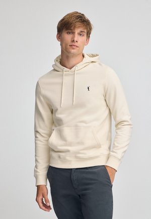 Haut en polaire crème avec poche kangourou, capuche avec cordon, et un petit logo noir sur la poitrine, porté avec un pantalon gris foncé.