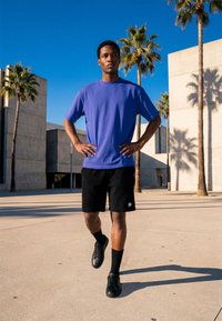 Homme en t-shirt bleu et shorts noirs marchant en plein air près de palmiers et de bâtiments modernes en béton sous un ciel bleu clair.