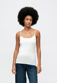 Kvinne med afrofrisyre iført en hvit ermeløs topp og mørkeblå jeans som står mot en enkel lyse bakgrunn.