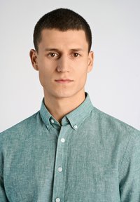 Ljusgrön skjorta med knappar och en texturerad tygkvalitet samt krage. Har vita knappar längs den främre plösen. Inga mönster.