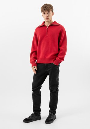 Jeune homme debout, portant un pull rouge à fermeture éclair sur le devant, un jean noir et des baskets noires, sur un fond blanc uni.