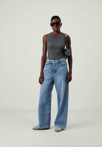 Grå linne, vida ljusblå jeans med dragsnöre, grå loafers och överdimensionerade mörka solglasögon. Grön mönstrad väska på axeln.