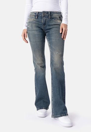Kvinde iført falmede blå bootcut-jeans med mindre slid og hvide sneakers, stående mod en ensfarvet baggrund.