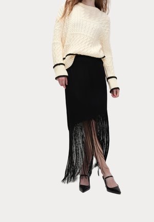 Femme portant un pull crème à torsades et une jupe noire avec de longues franges, associée à des talons noirs à bout pointu.