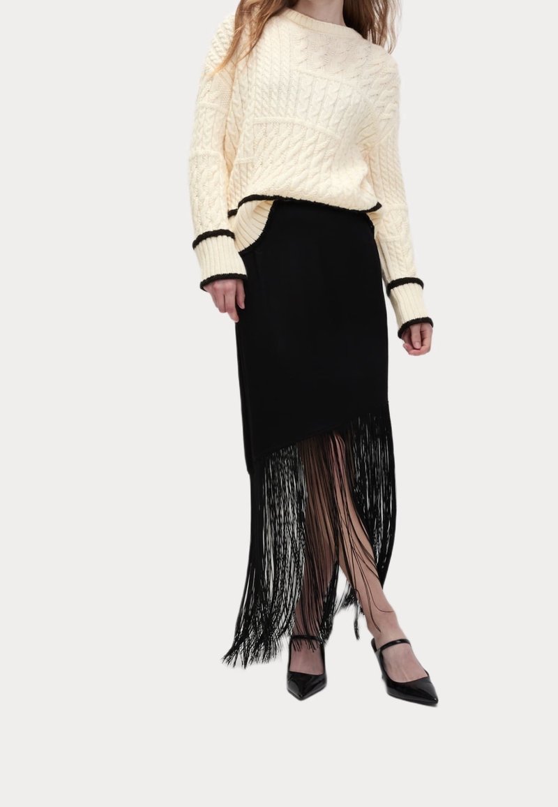Femme portant un pull crème à torsades et une jupe noire avec de longues franges, associée à des talons noirs à bout pointu.