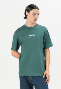 Zaļa īsroču t-krekls, izgatavots no kokvilnas, ar balto rakstaino "high tea" logo uz krūtīm, brīvās formas, vienkāršs dizains.