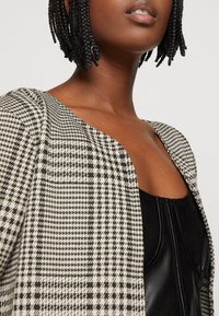 Veste à motif pied-de-poule noir et blanc, dotée d'un col arrondi et d'épaules structurées, portée sur un haut noir.