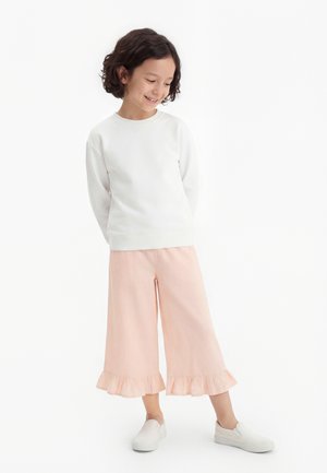 Pige med kort krøllet hår iført hvid langærmet skjorte, lyserøde vide bukser med flæser og hvide slip-on sko, stående og smilende.