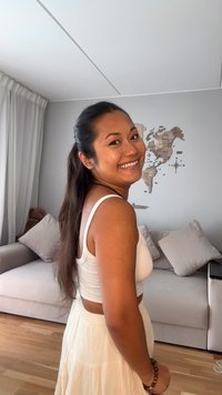 Weißes Tanktop und fließender weißer Rock, lange dunkle Haare, steht in einem Wohnbereich mit einem hellen Sofa und einer Wanddekoration mit einer Karte.