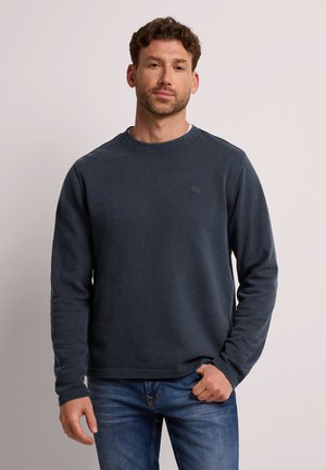 Uomo con capelli corti e ricci che indossa una felpa a maniche lunghe blu scuro e jeans blu, in piedi davanti a uno sfondo chiaro e uniforme.