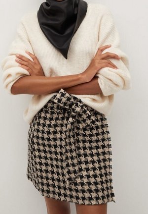 Gestrickter cremefarbener Pullover mit übergroßen Ärmeln, kombiniert mit einem schwarz-beigen Hahnentrittrock, der eine vordere Schleifendetail aufweist.