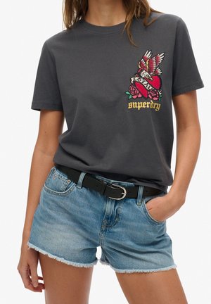Szara bawełniana koszulka z żywym haftowanym wzorem przedstawiającym serce, róże i tekst. Połączona z jasnoniebieskimi dżinsowymi szortami i czarnym paskiem.
