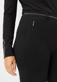 Zwarte broek gemaakt van rekbaar materiaal, met een zijzak met rits en een slim fit. De tailleband heeft een zilveren accentlijn.