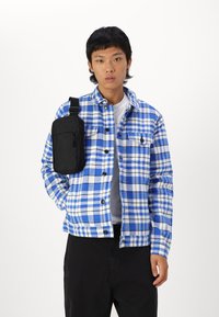 Blauw-wit geruite flanellen shirt met zwarte knopen en een borstzak, gecombineerd met een zwarte schoudertas. Zwarte broek zichtbaar.