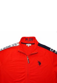 Röd zip-up jacka med hög krage, med marinblå accenter, broderad logotyp och vit text på axlarna. Materialet är slätt.