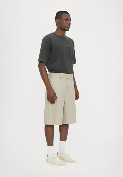T-shirt gris foncé à manches courtes, shorts beiges, chaussettes blanches et baskets claires. Détail de bouton notable sur les shorts, texture de tissu lisse.