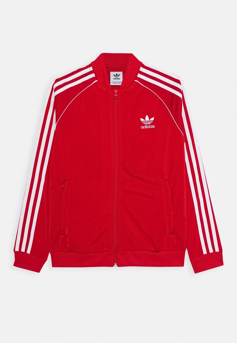 Veste de survêtement rouge à fermeture éclair fabriquée en tissu doux, avec des accents blancs à trois bandes sur les manches et un logo blanc sur la poitrine. Deux poches latérales.