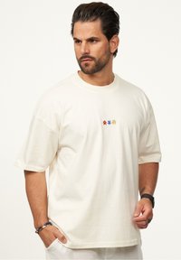 Cremfarbene Baumwoll-T-Shirt mit kurzen Ärmeln, verziert mit mehrfarbiger Stickerei auf der Brust. Lässiger Schnitt und glatte Textur.