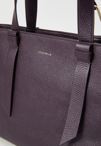 Purple textured leather tote bag with dual handles and a gold logo. Features flat straps and a structured shape, emphasizing simplicity.