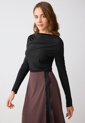 Donna con capelli castani a lunghezza spalle che indossa una maglia nera a maniche lunghe e una gonna marrone con dettagli in pizzo nero verticale.