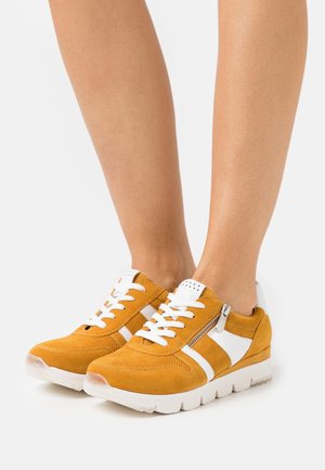 Des baskets en daim jaune présentent des accents et des lacets blancs, ainsi qu'une fermeture éclair sur le côté. Le design comprend une épaisse semelle blanche pour un confort supplémentaire.