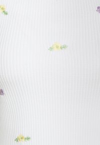 Vit ribbad textil med små broderade blommor i gult, grönt och lila, med en slät textur och figurnära design.