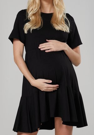 Pregnant woman with long blonde hair wearing a loose black dress, gently cradling her baby bump with both hands.